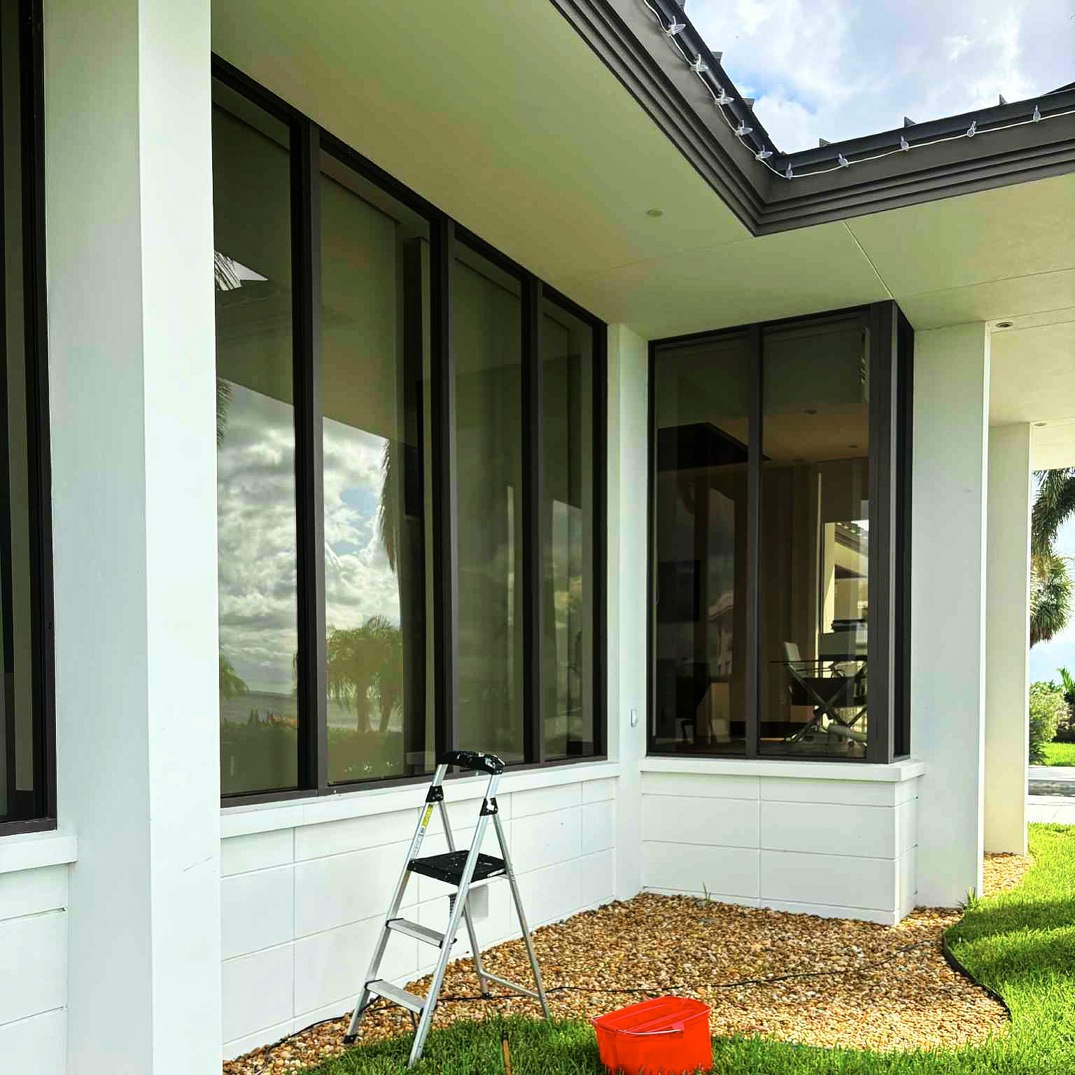 House exterior with black windows, a step ladder, red bucket, and blue towel on grass.